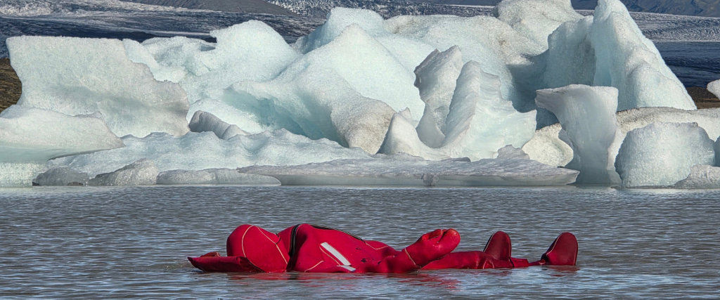 A picture showing a person floating on Fjallsárlón glacier lagoon in a thermal suit with icebergs in the background.